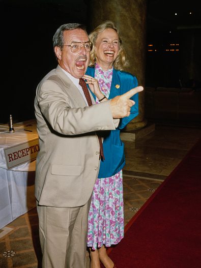 American actor William Daniels, wearing a beige suit, his mouth open as he wags his finger in mock anger, beside his laughing wife, American actress Bonnie Bartlett, who wears a blue jacket over a floral print dress, a sign reading 'Reception' behind them, United States, circa 1990. (Photo by Vinnie Zuffante/Getty Images)
